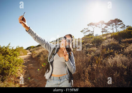 Felice ed energico giovane donna prendendo un selfie. Lei tiene la fotocamera del cellulare ad alta in posa e gesticolando vittoria il segno w