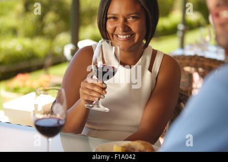 Ritratto di giovane e bella donna seduta a un tavolo in cantina con in mano un bicchiere di vino rosso e sorridente alla fotocamera. Foto Stock