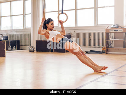 Montare la giovane donna facendo pull-up su anelli di ginnastica. Muscoloso giovane atleta femminile esercizio con anelli in palestra. Foto Stock
