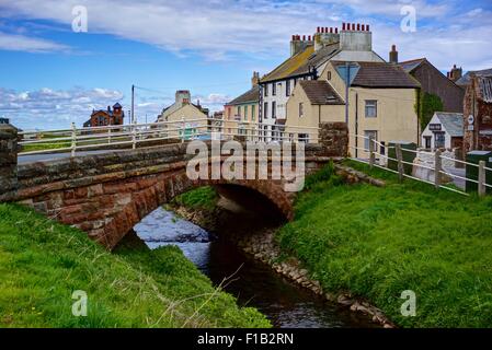 In rosso di un ponte in pietra che attraversa il fiume Ellen in Alonby, Cumbria, Inghilterra., con un assortimento di case dipinte, cielo blu, verde erba. Foto Stock