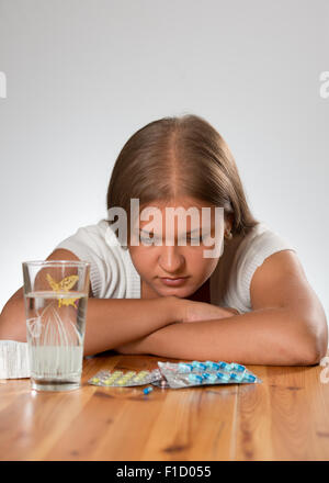 Triste giovane donna seduta sul tavolo sullo sfondo bianco con alcune pillole e bicchiere di acqua Foto Stock