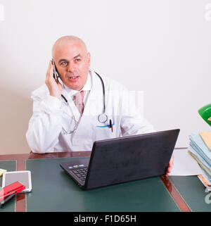 Sconcertato medico nel suo studio, utilizza uno smartphone davanti al suo computer portatile. Utilizzare le nuove tecnologie. Nel suo studio professionale, egli è si Foto Stock