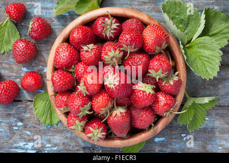 Fragole mature nella ciotola di legno e foglie verdi sulla vecchia tabella, vista dall'alto. Foto Stock