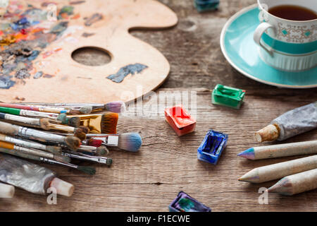 Pennelli, artista tavolozza, matite, tazza di caffè, acquarello e colori a olio sulla scrivania in studio di pittore. Foto Stock