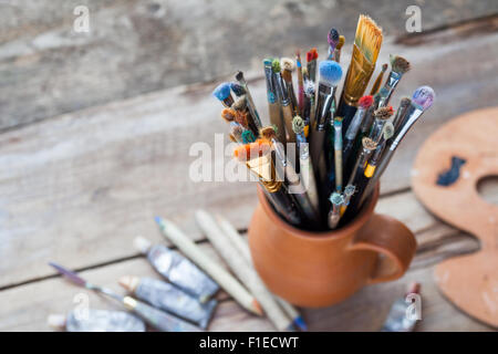 Pennelli in una brocca da potters argilla, tavolozza e tubi di vernice sulla scrivania. Foto Stock
