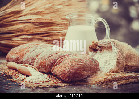 Pane fresco, brocca del latte, sacco di farina di grano e un covone di grano su un tavolo di legno. Retrò stilizzato. Foto Stock