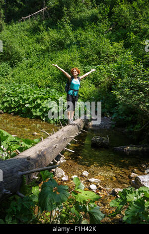 Felice escursionista femmina che attraversa un fiume di montagna su un enorme tronco di albero Foto Stock
