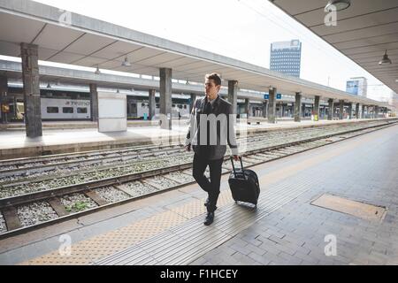 Giovane imprenditore commuter percorrendo a piedi Stazione ferroviaria piattaforma tirando la valigia. Foto Stock