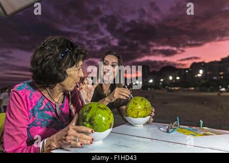 Donna matura e madre di bere da gusci di noce di cocco in spiaggia di notte, Copacabana, Rio de Janeiro, Brasile Foto Stock