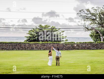 Vista posteriore della sposa e lo sposo tenendo le mani alzate in giardino di Hawaiian wedding, Kaaawa, Oahu, Hawaii, STATI UNITI D'AMERICA Foto Stock