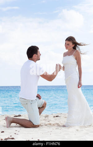 Uomo in giù su un ginocchio e chiedendo per sposare la sua donna, tenendo la mano sulla spiaggia di sabbia vicino al mare per una vacanza nei Caraibi. Foto Stock