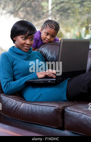 Giovane africana Madre giace su un lettino mentre si lavora su un computer portatile, sua figlia guardando sopra la sua spalla Foto Stock