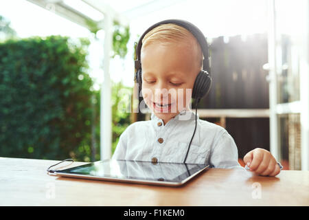 Felice di ridere little boy ascolta musica sul suo tablet pc attraverso le cuffie stereo come egli siede all'aperto in una calda giornata estiva Foto Stock