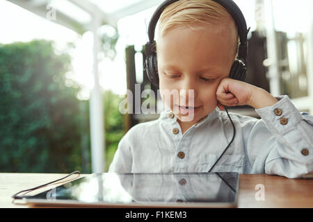 Adorable del giovane ragazzo seduto con un computer tablet ascoltando la sua libreria musicale con un sorriso di contentezza su un set di s Foto Stock