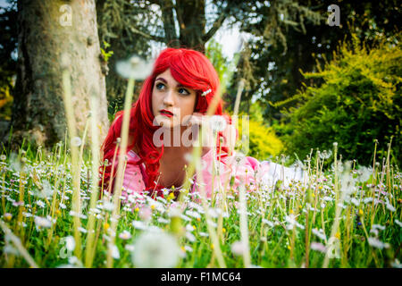 Giovane donna con luminosi capelli rossi sdraiato nella foresta di Campo dei Fiori con la mano appoggiata sul mento cercando Dreamily Off nello spazio Foto Stock