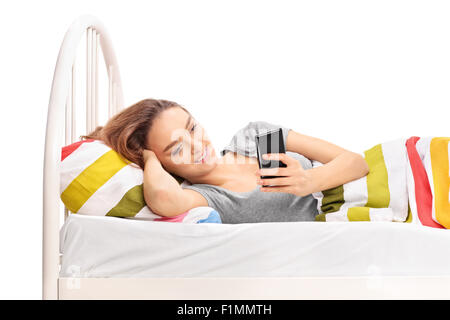 Bruna giovane donna giaceva a letto e guardando qualcosa sul suo telefono cellulare isolato su sfondo bianco Foto Stock