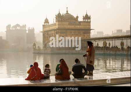 Amritsar Punjab, India. Il Tempio d'Oro - Harmandir Sahib - all'alba con un vecchio uomo Sikh saluto una famiglia seduta con le gambe incrociate sul lato del lago. Foto Stock