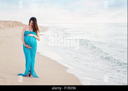 Donna incinta su una spiaggia indossando un turchese blu abito di maternità. Lei è sempre sorridente e guardando verso il basso al suo ventre. Foto Stock