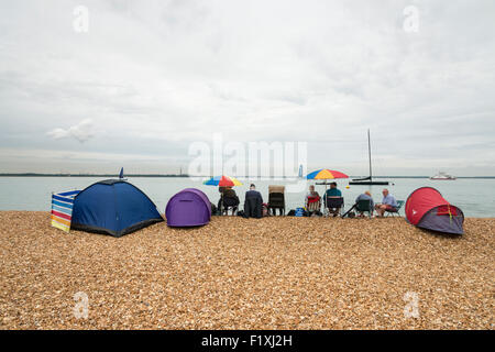 La gente seduta sulla spiaggia sotto gli ombrelloni in nuvoloso meteo guarda il racing in Cowes Week, Isle of Wight REGNO UNITO Foto Stock