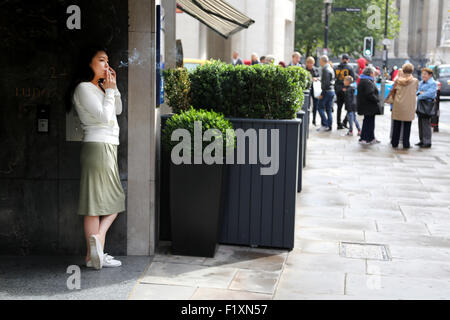 Londra 2015: un lavoratore di ufficio prende una pausa sigaretta al di fuori del suo ufficio edificio quali fronti su una strada trafficata. Lei con il suo mobile Foto Stock