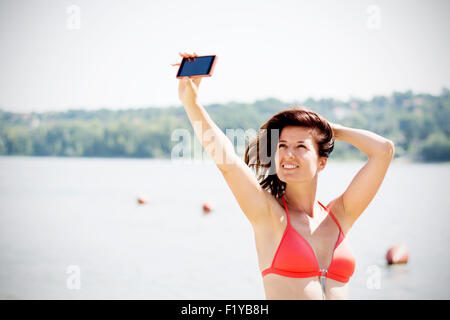 Attraente bruna giovane donna prendendo autoritratto con il suo telefono sulla spiaggia. Indossa red bikini e sorridente. Foto Stock