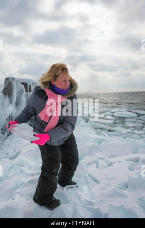 Donna bionda getta dei pezzi di ghiaccio da congelato sulla riva lago superiore nel nord del Minnesota su una giornata invernale Foto Stock