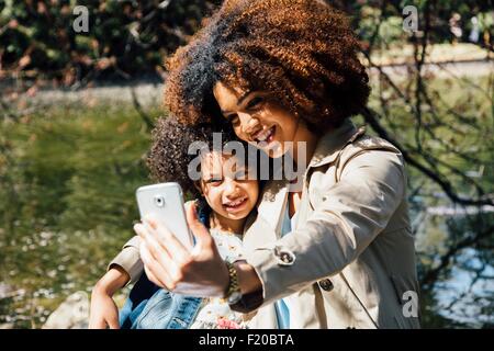 Madre con braccio attorno alla figlia utilizza lo smartphone per prendere selfie Foto Stock
