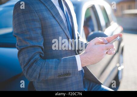 Ritagliato shot del giovane imprenditore appoggiata contro auto utilizza lo smartphone Foto Stock