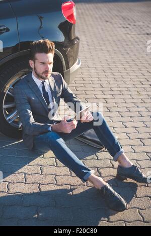 Elegante giovane uomo seduto appoggiato contro auto con tavoletta digitale Foto Stock