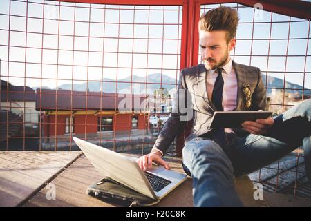Elegante giovane uomo seduto su passerella utilizzando laptop e tablet digitale Foto Stock