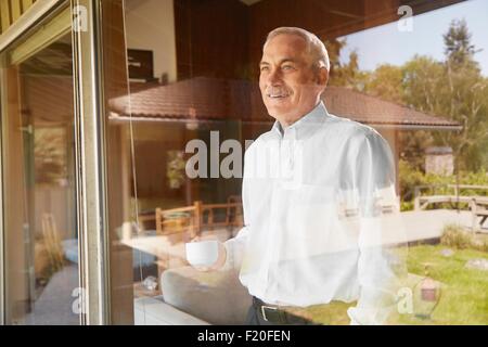 Senior uomo a casa e di bere caffè, guardando fuori della finestra Foto Stock