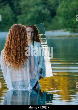 Giovane donna in piedi nel lago, tenendo specchio, guardando la riflessione Foto Stock