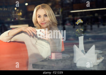 Molto bella giovane donna, sedersi al bar e bere un tè o un caffè con croissant, street la visuale della finestra frontale Foto Stock