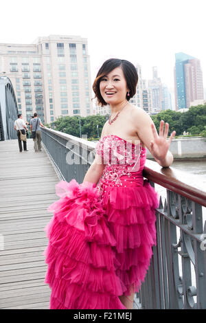 Felice sposa cinesi indossando un moderno rosa abito da sposa le onde a telecamera come essa si erge sul Bund bridge, Shanghai, Cina e Asia Foto Stock