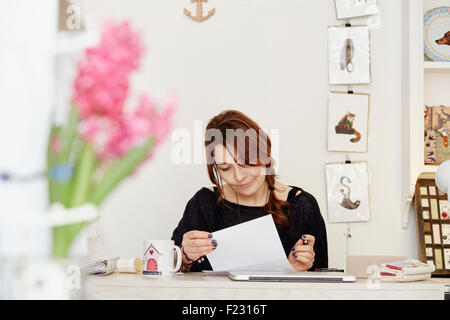 Una donna seduta a una scrivania in un piccolo negozio di articoli da regalo, facendo i documenti, la gestione del business. Un laptop sulla scrivania. Foto Stock