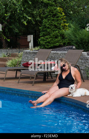 Rivestimento donna caucasica nel suo 50s si siede sul bordo di una zona residenziale piscina nel suo bikini petting il suo piccolo cane bianco Foto Stock