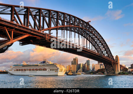 Oversized ocean liner crociera passando da sotto il Ponte del Porto di Sydney al Tramonto backgrounded dai grattacieli della città Foto Stock