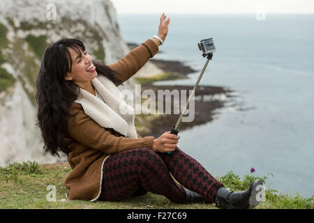 Giovane donna prendendo un selfie con una GoPro camera e un bastone selfie. Le Bianche Scogliere di Dover, Dover, Kent, Regno Unito Foto Stock