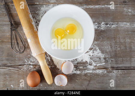 La cottura di sfondo con uova, uova crude in un piatto da portata, gusci d'uovo, la farina, il mattarello e frusta su uno sfondo di legno. Foto Stock