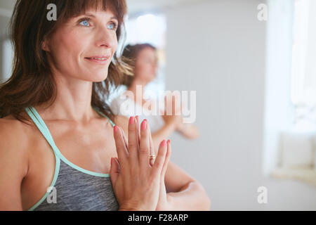 Immagine ravvicinata di felice donna matura fare yoga che guarda lontano sorridente. Le donne in posa yoga vrikshasana utilizzando Namaste. Foto Stock