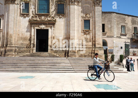 Una signora in bicicletta attraverso una piazza davanti a una chiesa. Foto Stock