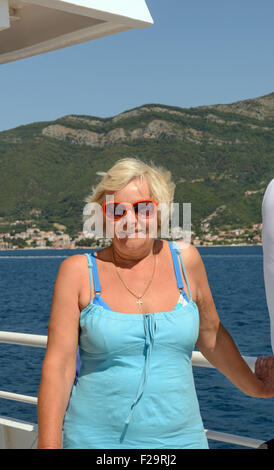 Senior bionda abbronzata donna sorridente è sul ponte dello yacht sulla costiera sullo sfondo di montagna, Kotor Bay, Montenegro. Foto Stock