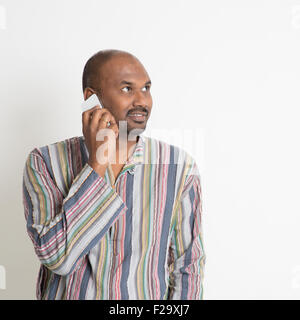 Ritratto di maturo business casual uomo indiano chiamando sullo smartphone, in piedi su sfondo semplice con l'ombra. Foto Stock