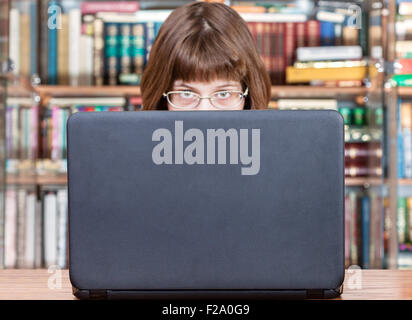 Ragazza con occhiali guarda al di sopra del coperchio del computer portatile aperto nella libreria Foto Stock