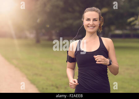 Mezzo Corpo colpo di una bella donna atletica Jogging al parco con le cuffie e sorridente alla fotocamera, con spazio di copia Foto Stock