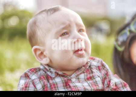 L'innocente la felicità di un simpatico 6 mesi baby con luce marrone capelli in rosso a scacchi maglietta e pantaloni beige sorridente in un parco della città Foto Stock