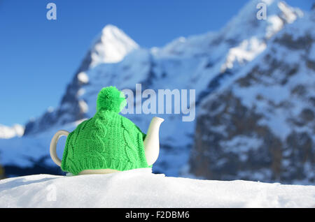 Tè pentola nel cappuccio di maglia sulla neve contro il picco di montagna Foto Stock