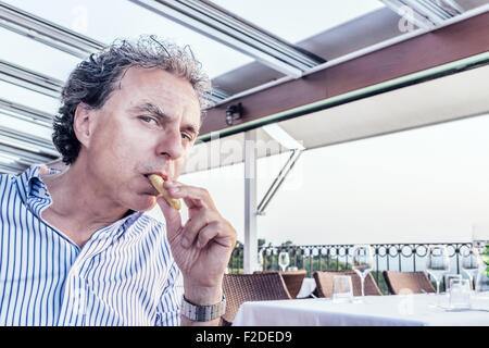 Momenti magici da condividere come una tranquilla cena presso il ristorante che si affaccia sul mare al tramonto: funny uomo di mezza età con la pelle morbida è mangiare pane stick Foto Stock