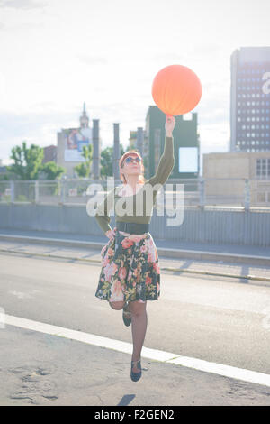 Giovani caucasici bello redhead donna jumping, giocando con palloncino arancione, mano sui suoi fianchi, cercando - gioventù, concetto di infanzia - indossare gonna floreale e camicia verde Foto Stock