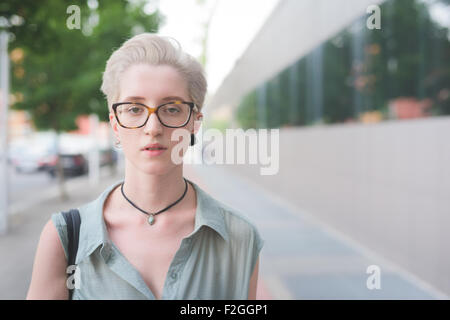 Ritratto di una giovane bella bionda caucasica designer italiano in posa nella città, cercando nella fotocamera, grave - indossando un screziato paio di occhiali e un vestito azzurro - gravità, pensieroso concept Foto Stock
